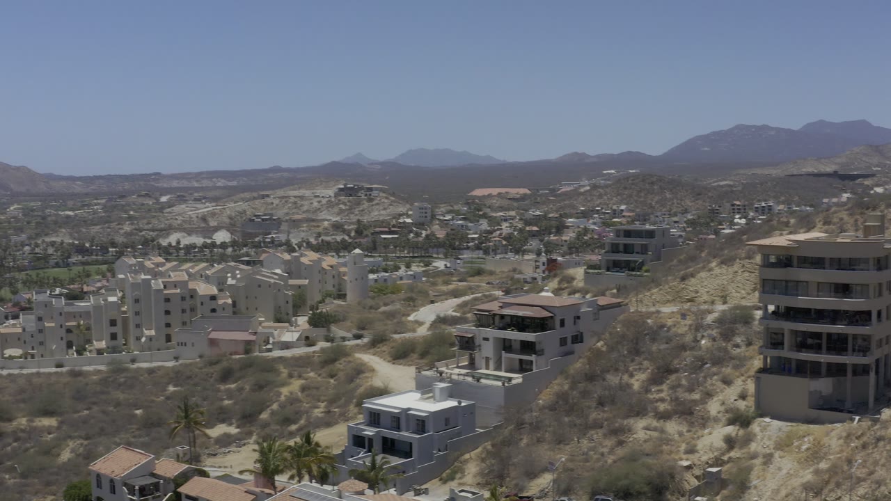 Aerial view in Los Cabos showing a modern boxy home near the lower frame, pale multi-story buildings on the hill, winding roads, and no visible people.