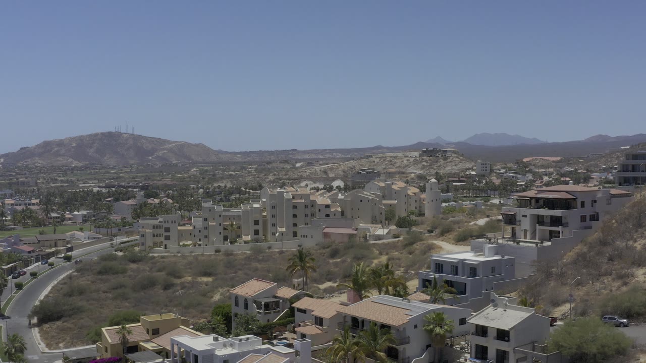 Aerial frame of Los Cabos, Mexico, with pale multi-story buildings in the midground, a curved road at left, dry hills, and no visible people.