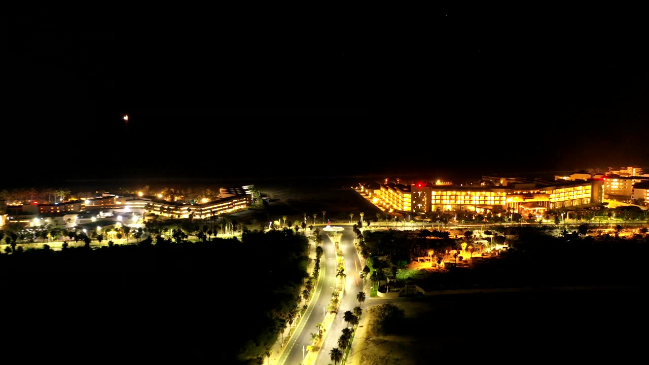 Night drone frame over Los Cabos, Mexico, with bright road lights leading toward large illuminated buildings and dark water beyond, with no visible people.