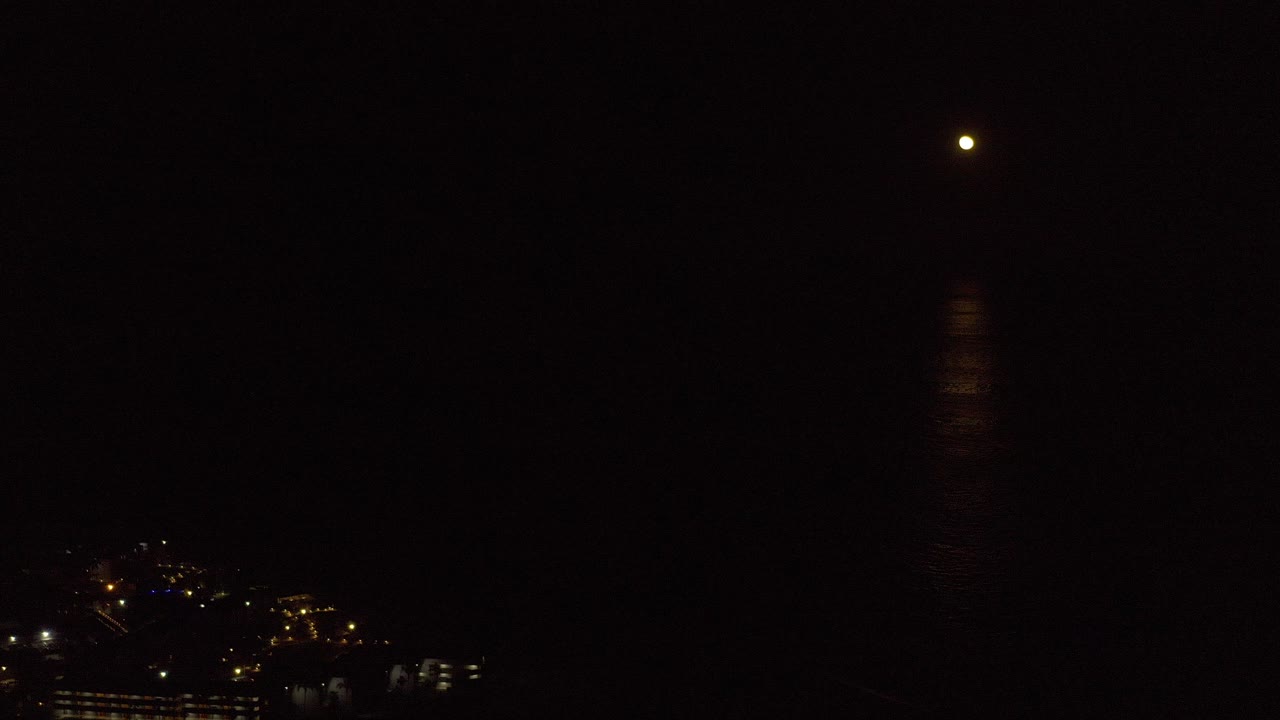 Opening frame of a Los Cabos night sea view with the moon above a faint vertical reflection and distant warm shoreline lights, no visible people.