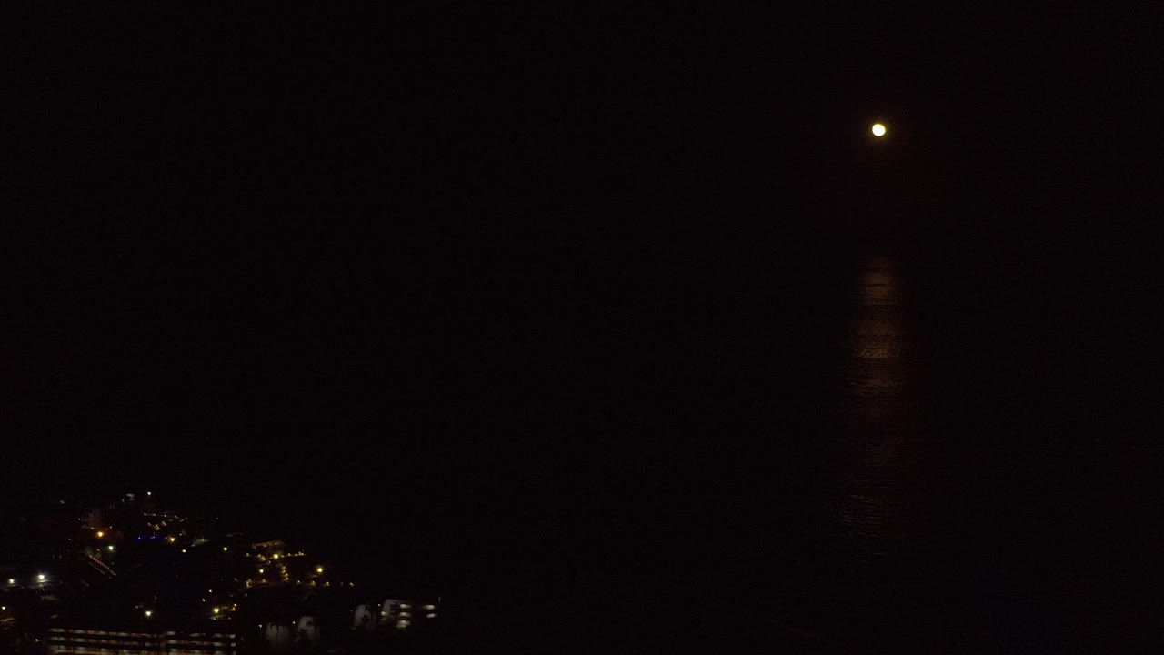 A Los Cabos night frame shows the moon reflection slightly clearer on the dark sea, with clustered city lights low on the left and no visible people.