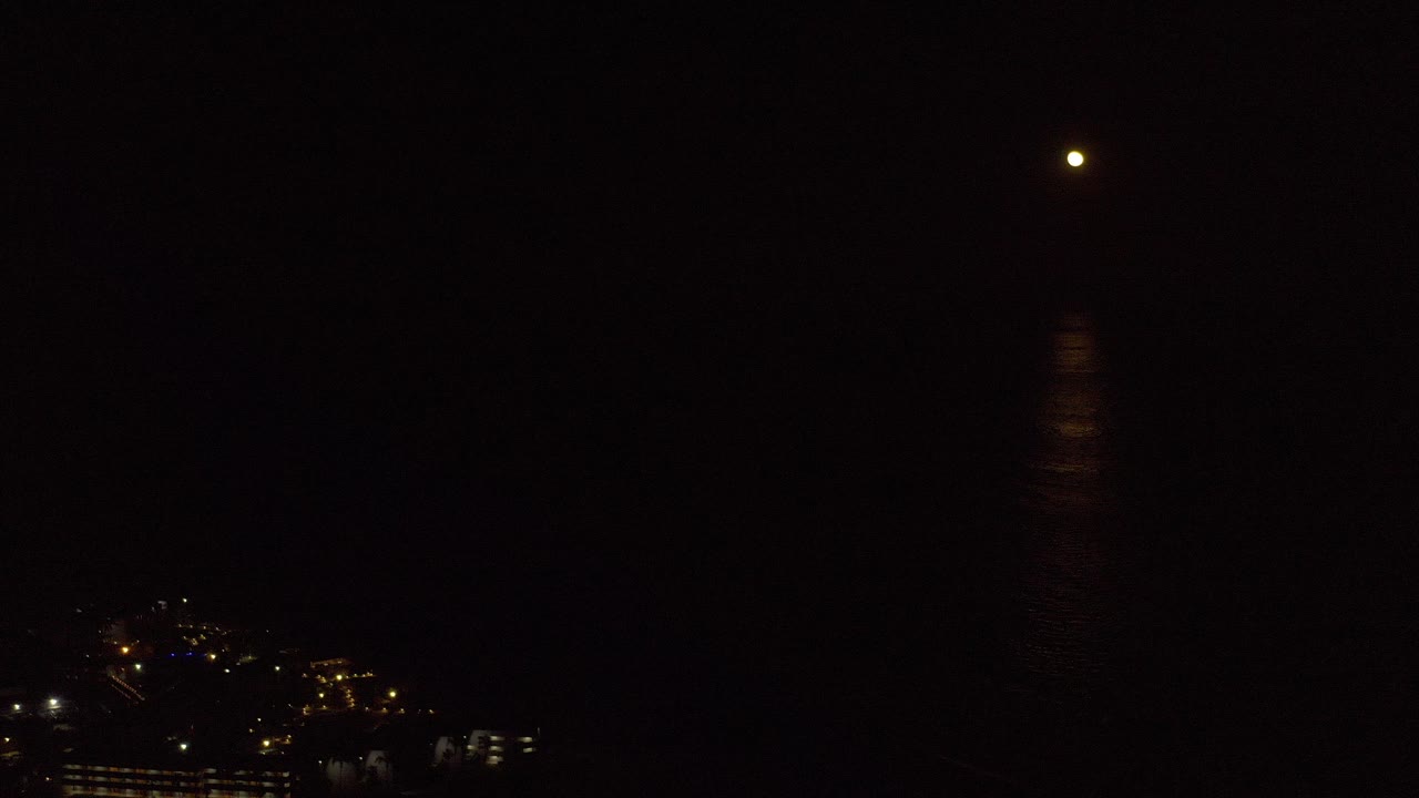 Small moon hangs over dark water in Los Cabos, Mexico, with a faint vertical reflection and distant shoreline lights at the lower left, no visible people.