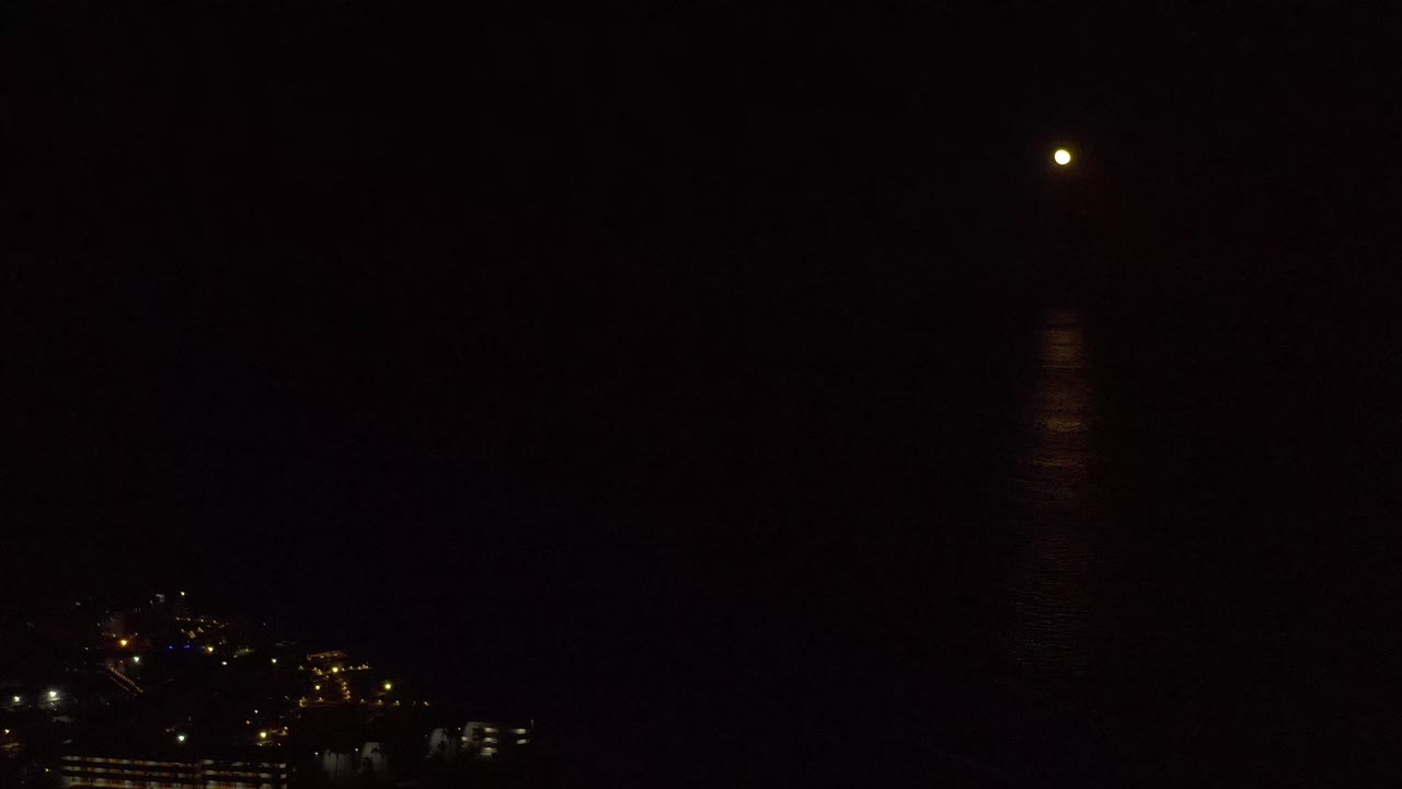 Moon and its muted reflection remain centered above dark water in Los Cabos, Mexico, while small clusters of city lights sit low on the left, no visible people.