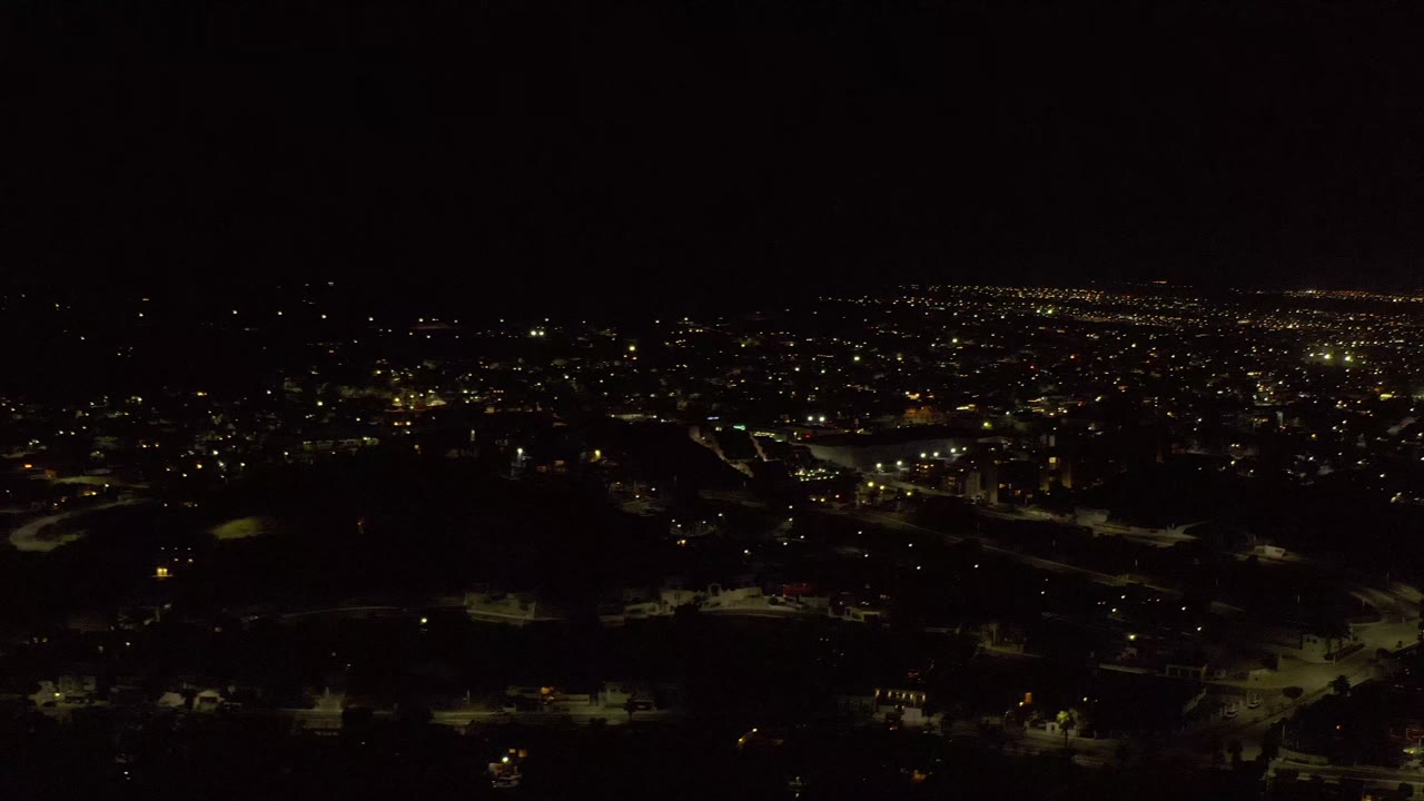 Night aerial frame in Los Cabos, Mexico showing dark foreground homes, warm streetlights, and a dense band of lights on the horizon, with no visible people.