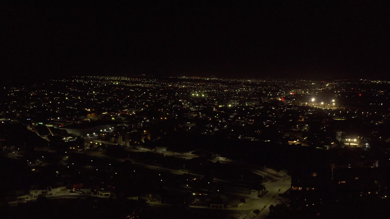 Wide elevated night view over Los Cabos, Mexico, with scattered city lights, a dark foreground hillside, bright distant light clusters, and no visible people.