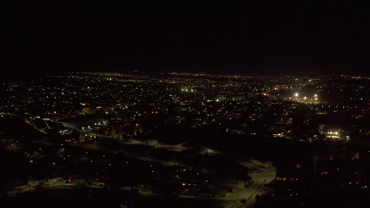 Elevated Los Cabos night frame showing a dense spread of small yellow city lights, dark lower foreground, brighter lights near the center-right horizon, and no visible people.