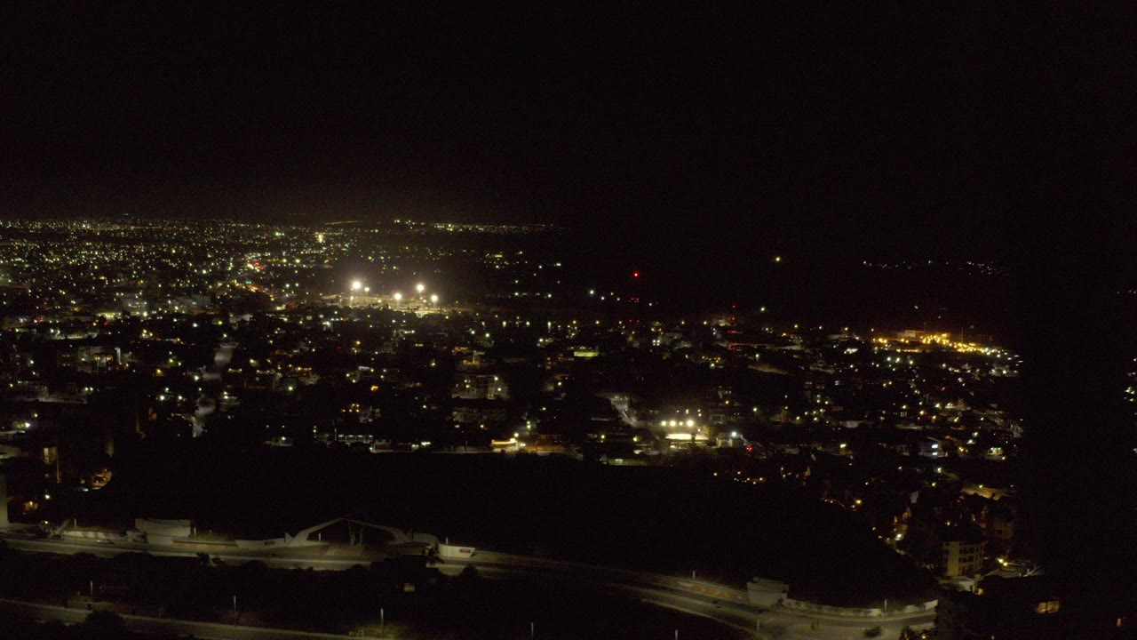 Opening frame of Los Cabos at night from an elevated aerial view, with dense city lights, dark water or shoreline in the foreground, and no visible people.
