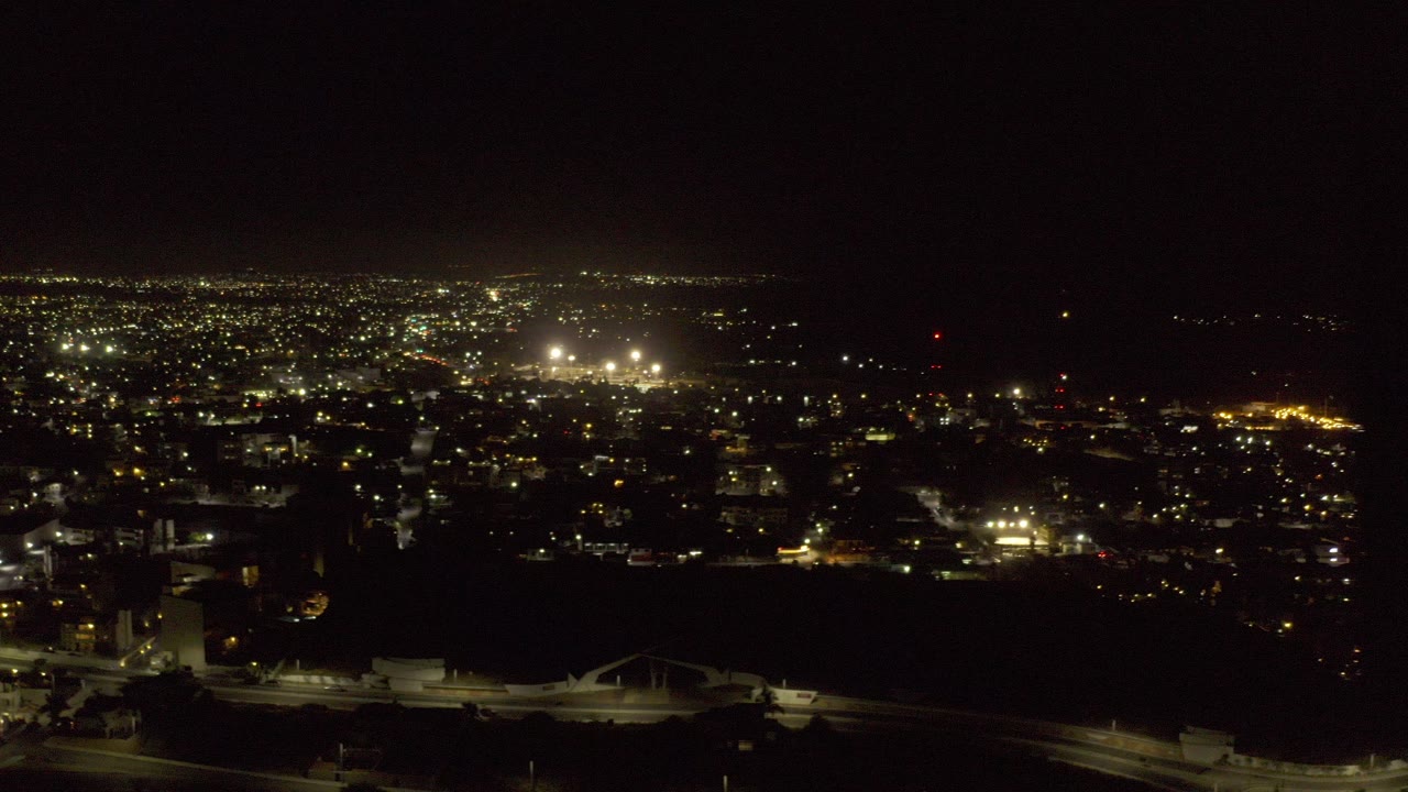 Opening frame of Los Cabos, Mexico at night from the air, with a dark foreground, scattered road lights, a bright city grid in the distance, and no visible people.