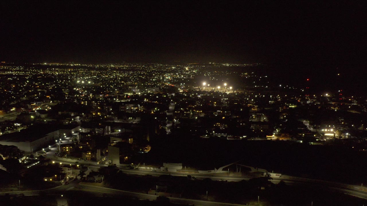 Early frame of a Los Cabos night aerial showing scattered city lights, dark foreground land, lit roads, low buildings, and no visible people.