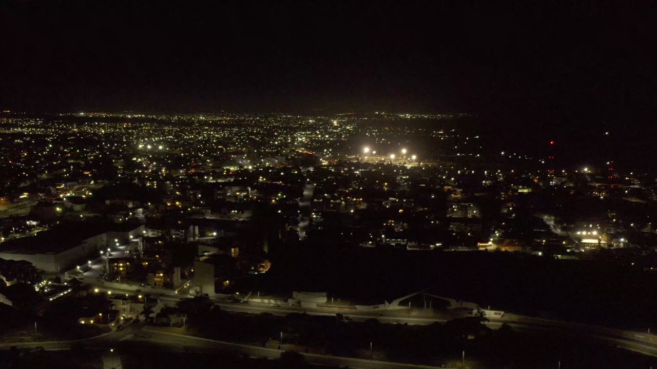 Los Cabos night drone frame with more lit building blocks visible below the horizon, a dark open foreground, and no visible people.