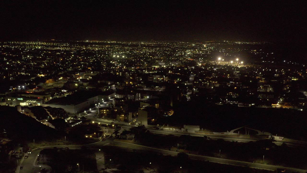 Night aerial view over Los Cabos, Mexico with lit roads, parking areas, low buildings, and a wide field of city lights on the horizon, with no visible people.