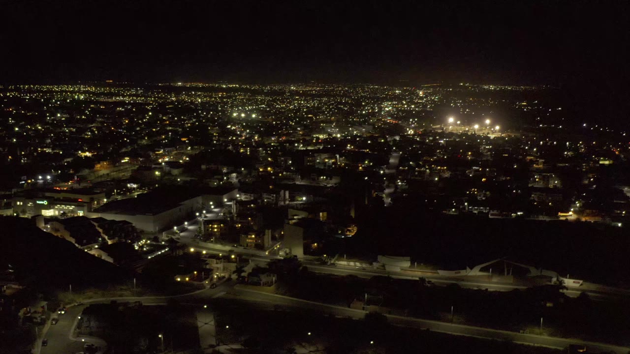 A second nighttime aerial frame of Los Cabos shows the same road network, lit building clusters, parking lots, and bright distant city lights, with no visible people.