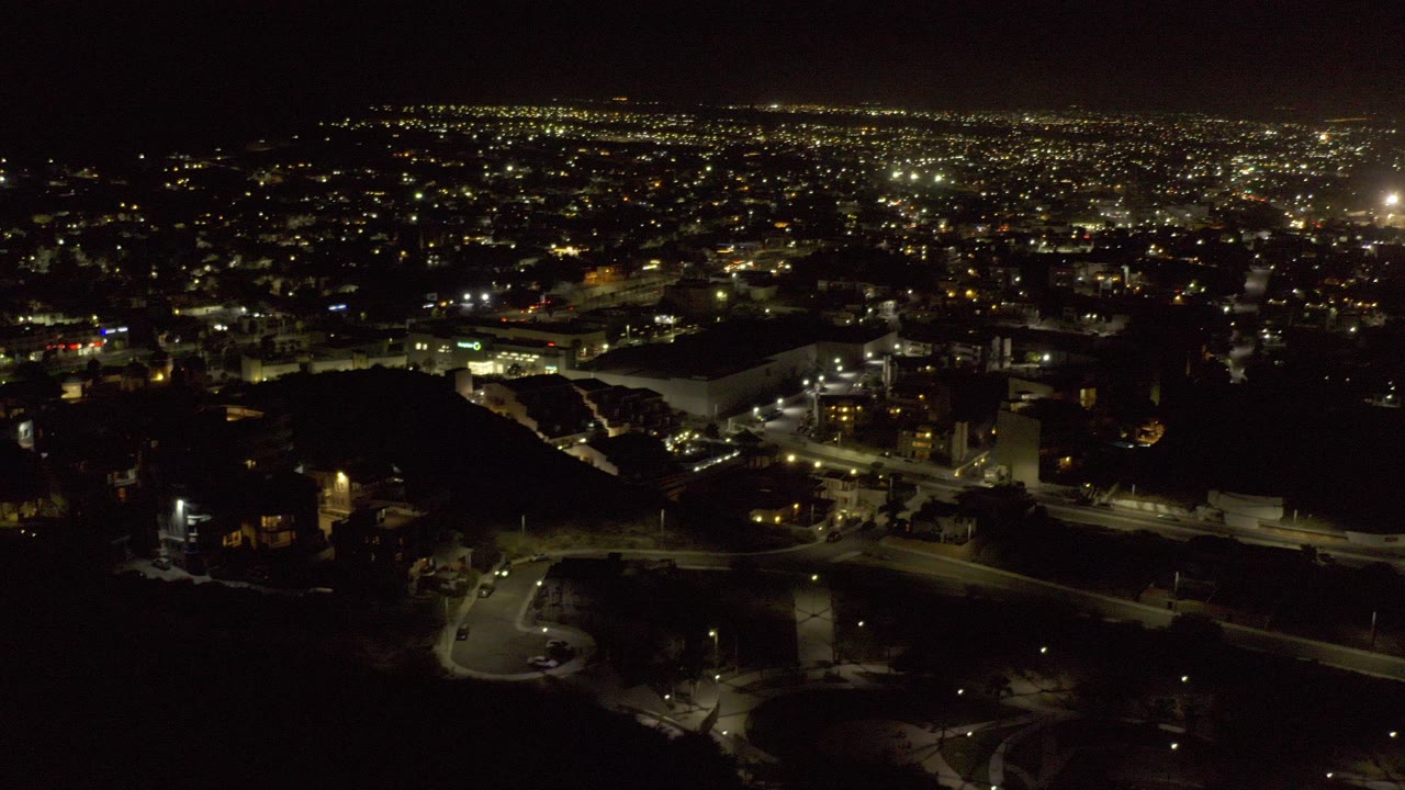 Opening frame of a Los Cabos, Mexico night aerial showing a broad field of city lights beyond dark foreground terrain, with roads and large buildings below and no visible people.