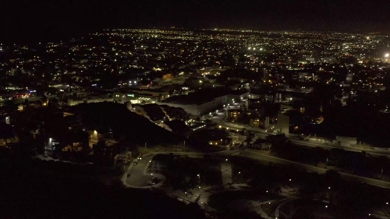 Second frame of the Los Cabos, Mexico aerial keeps the city-light horizon prominent while roads and blocky buildings sit in the darker lower portion, with no visible people.
