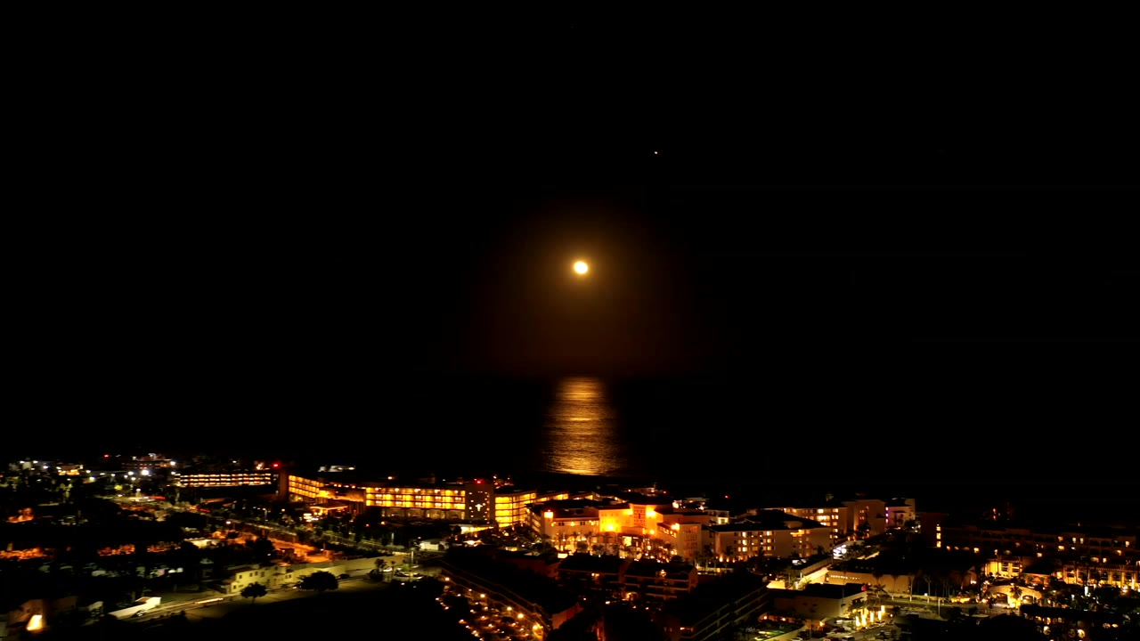 Full moon over dark ocean and illuminated waterfront buildings at night.
