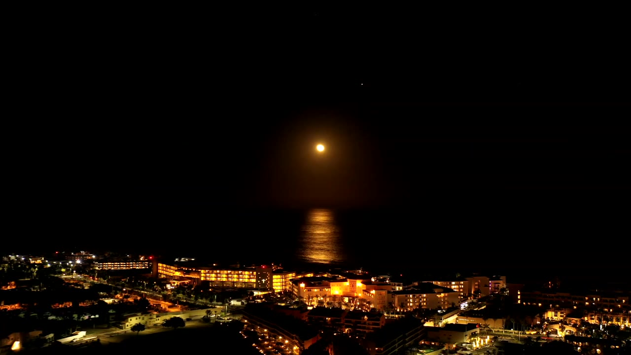 Los Cabos at night with the full moon slightly above the horizon, a vertical gold reflection on dark water, lit buildings below, and no visible people.