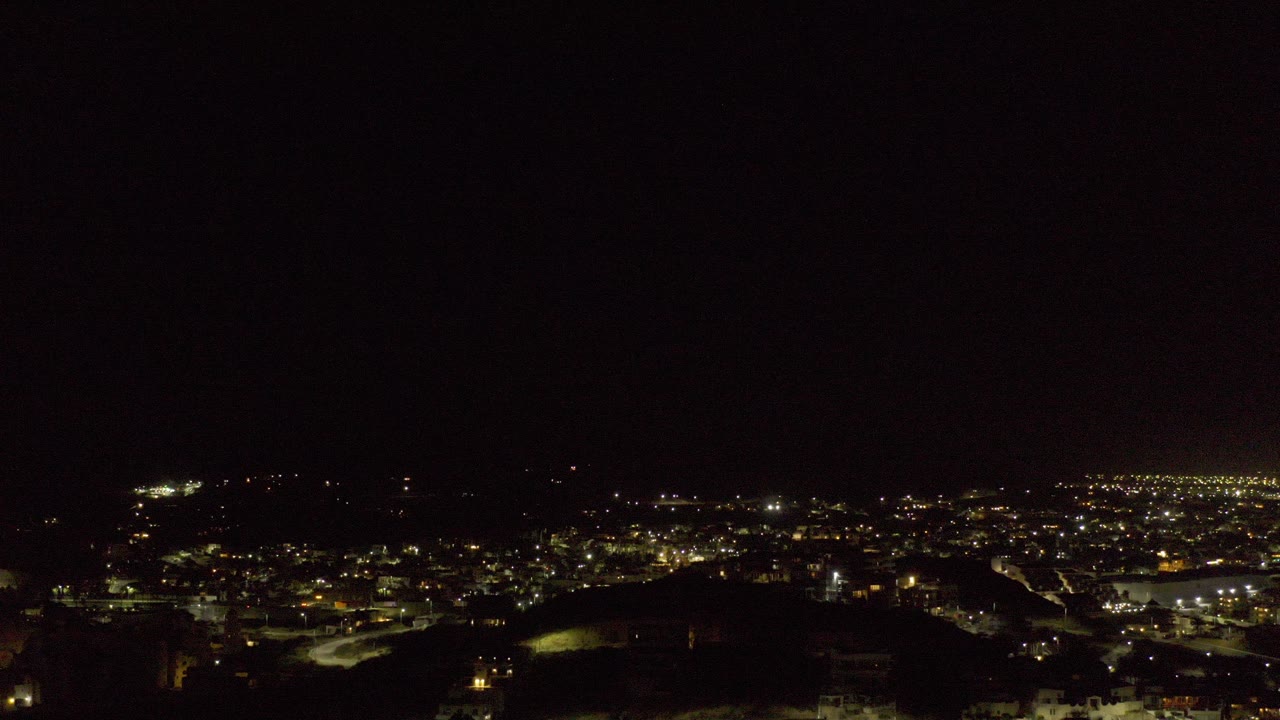 Opening frame of a dark Los Cabos night view with a broad black sky, a shadowed foreground ridge, distant city lights, and no visible people.