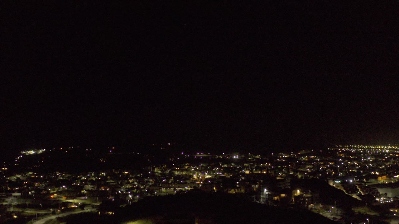 Opening view of Los Cabos at night from an elevated vantage, with a dark foreground slope, scattered city lights, a glowing horizon, and no visible people.