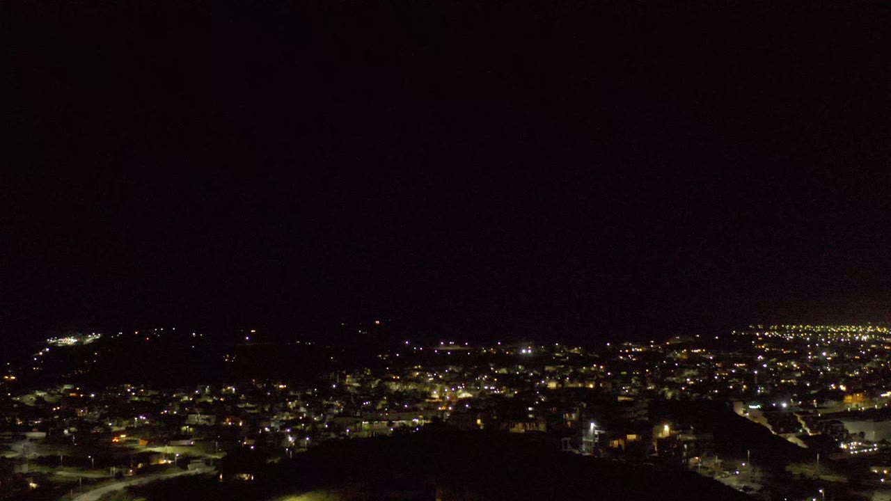Los Cabos city lights stretch across the frame under a mostly black sky, with the dark hillside still visible at the bottom and no visible people.