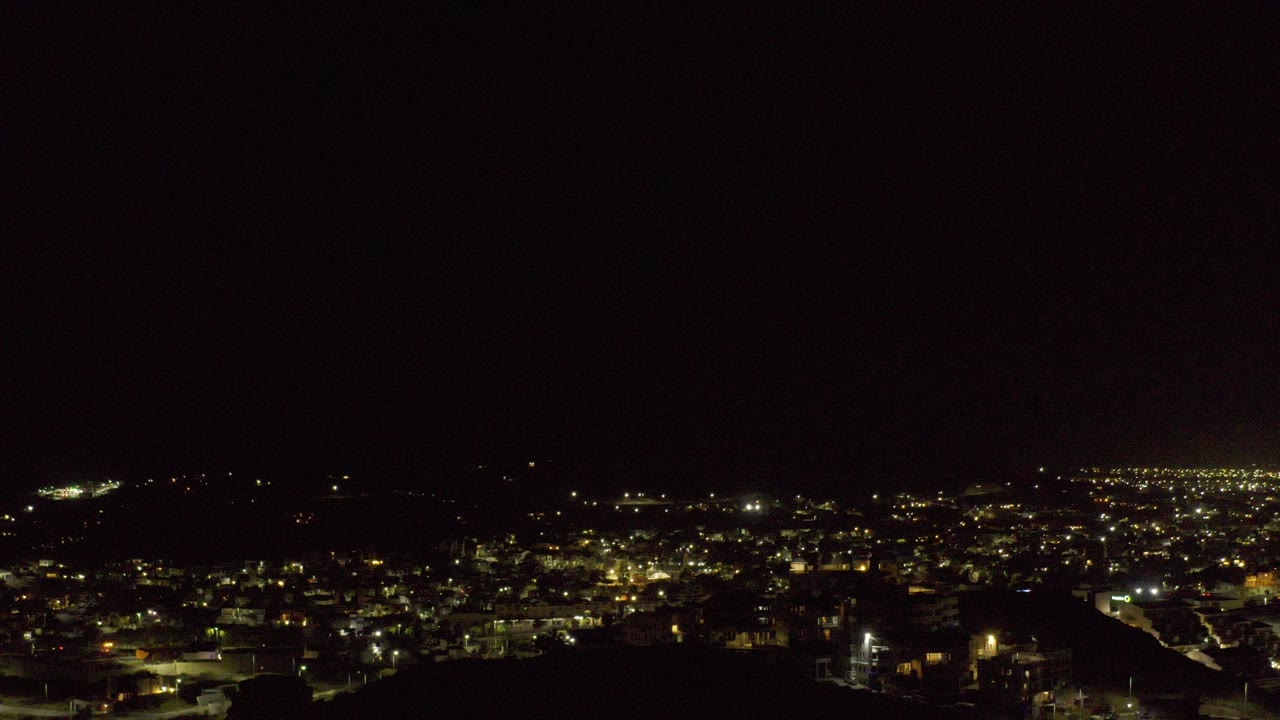 Wide nighttime view of Los Cabos, Mexico, with yellow city lights across the lower frame, dark hills in the distance, and no visible people.