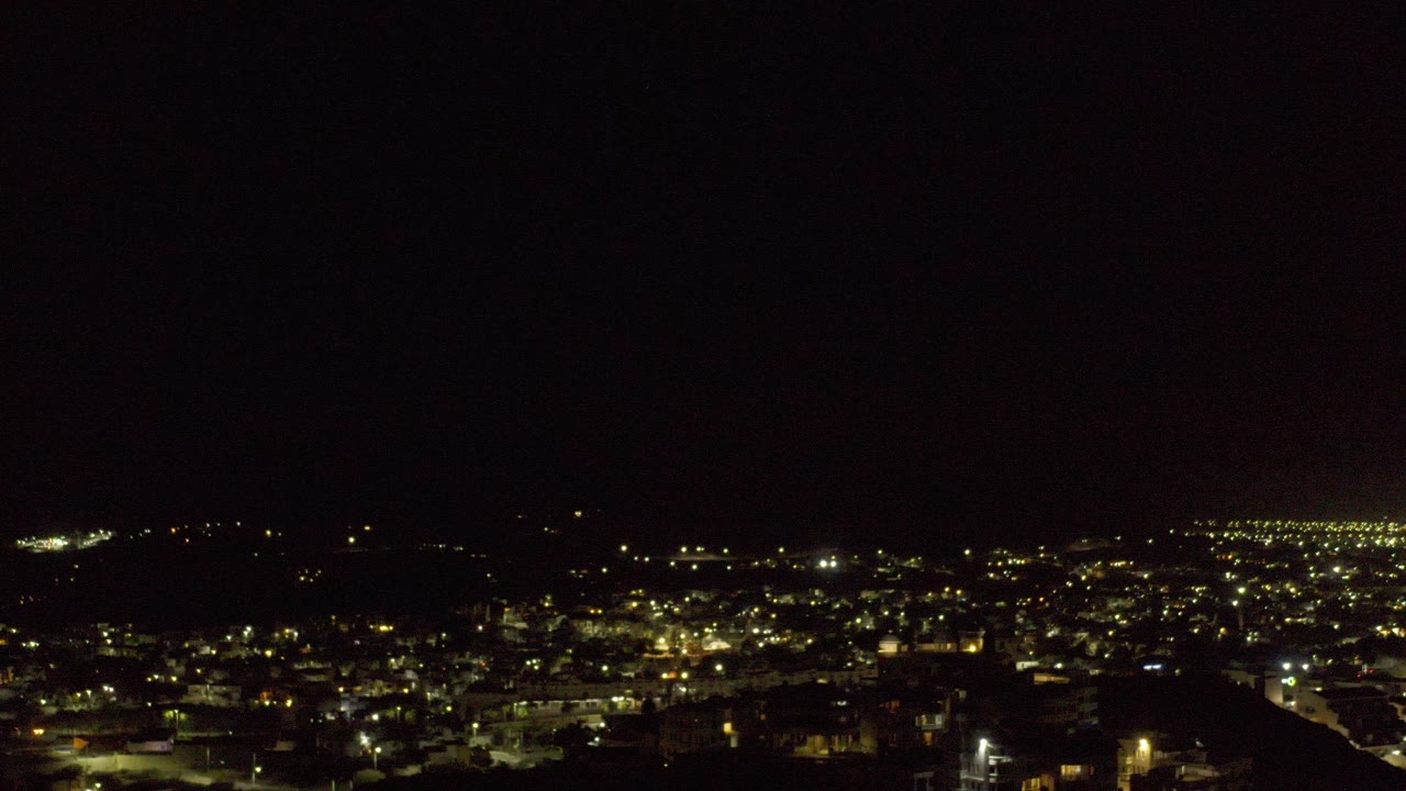 Wide night view of Los Cabos, Mexico with warm town lights across the horizon, a dark foreground slope, and no visible people.