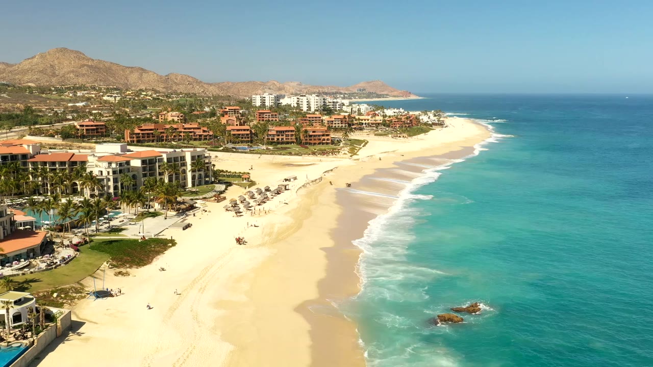 Los Cabos aerial frame with the shoreline running diagonally past resort hotels, pale sand, low surf, beach umbrellas, tiny people, and hills under clear light.