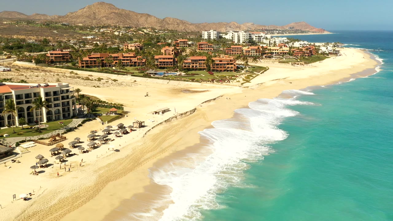 Aerial frame over Los Cabos, Mexico showing a golden beach beside turquoise water, resort buildings near the shore, dry hills inland, and no visible people.