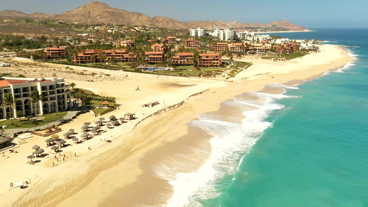 Aerial view in Los Cabos, Mexico with white waves curling onto a wide sand beach, red-toned resort buildings inland, blue water at right, and no visible people.