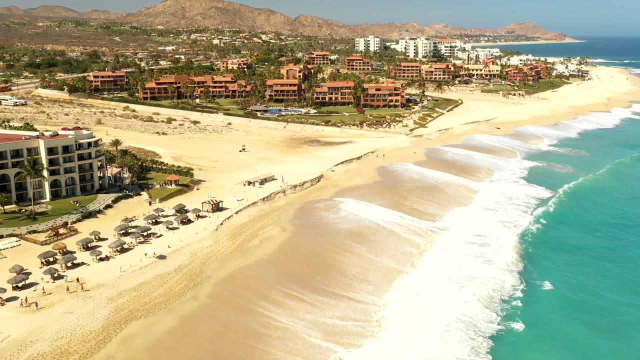Aerial coastline view with waves, beach umbrellas, resorts, and hills.