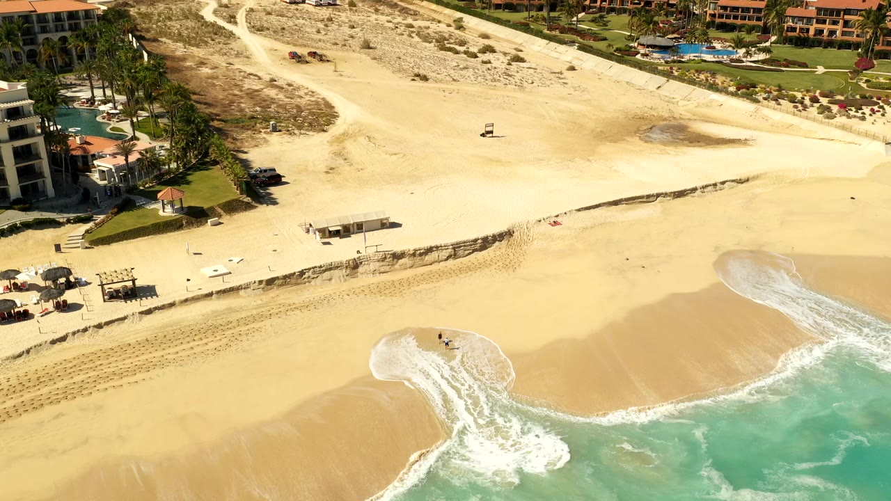 Aerial view of sandy beach, resort buildings, palms, and surf.