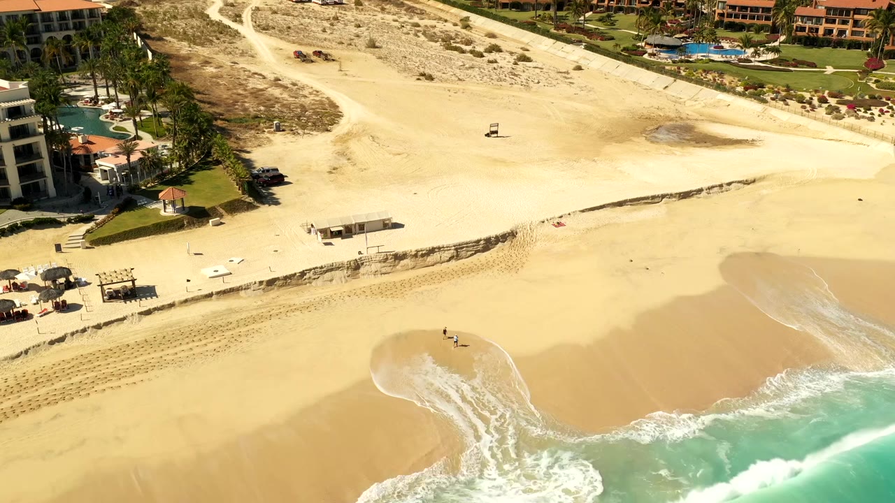 Los Cabos shoreline from above with a rounded patch of wet sand near the surf, resort buildings and palms above, and tiny figures visible on the open beach.