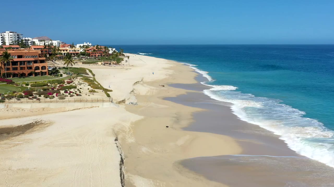Early frame of the Los Cabos shoreline with white surf edging the sand, clustered resort buildings at upper left, and no visible people on the beach.