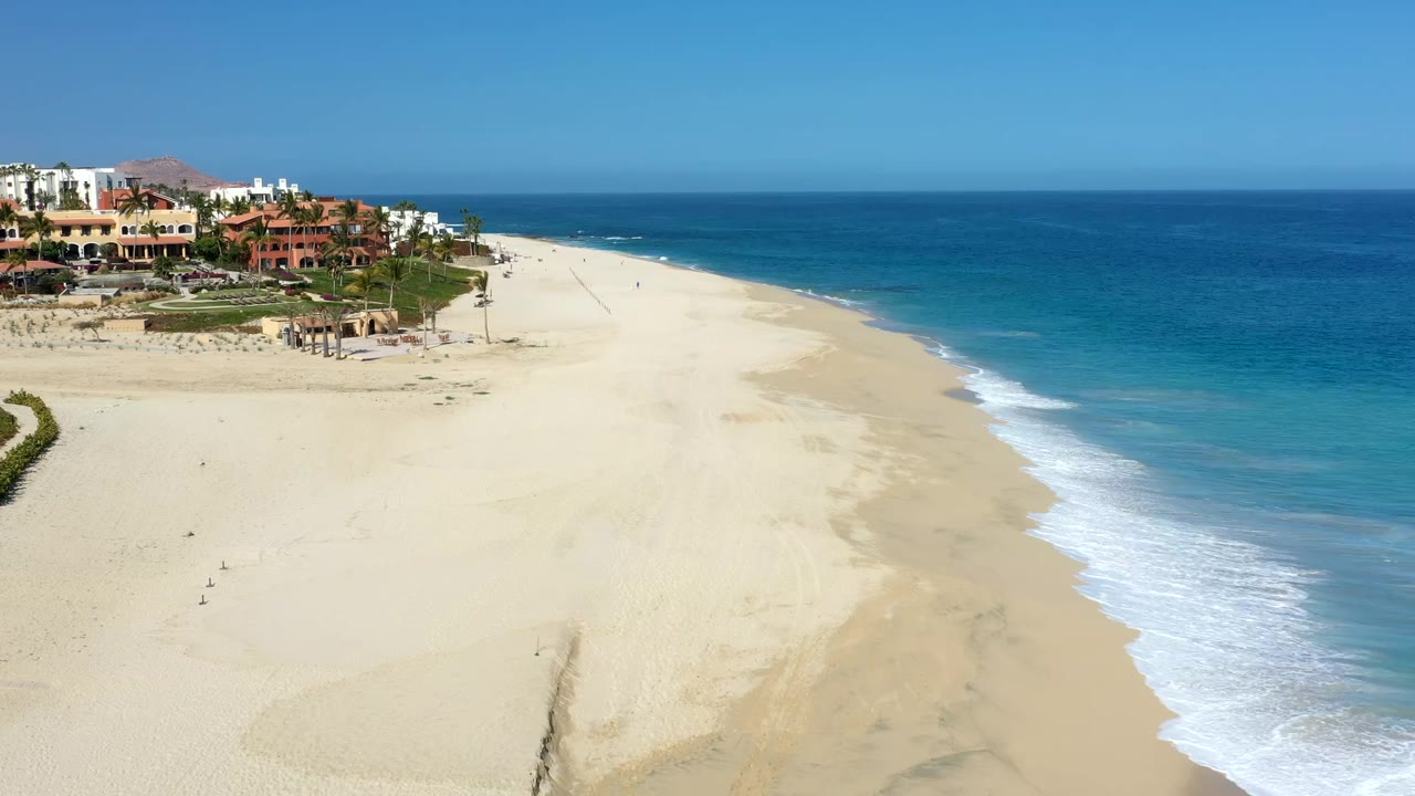 Los Cabos aerial view with the sandy beach filling the lower frame, turquoise shallows along the surf line, deeper blue water beyond, and no visible people.