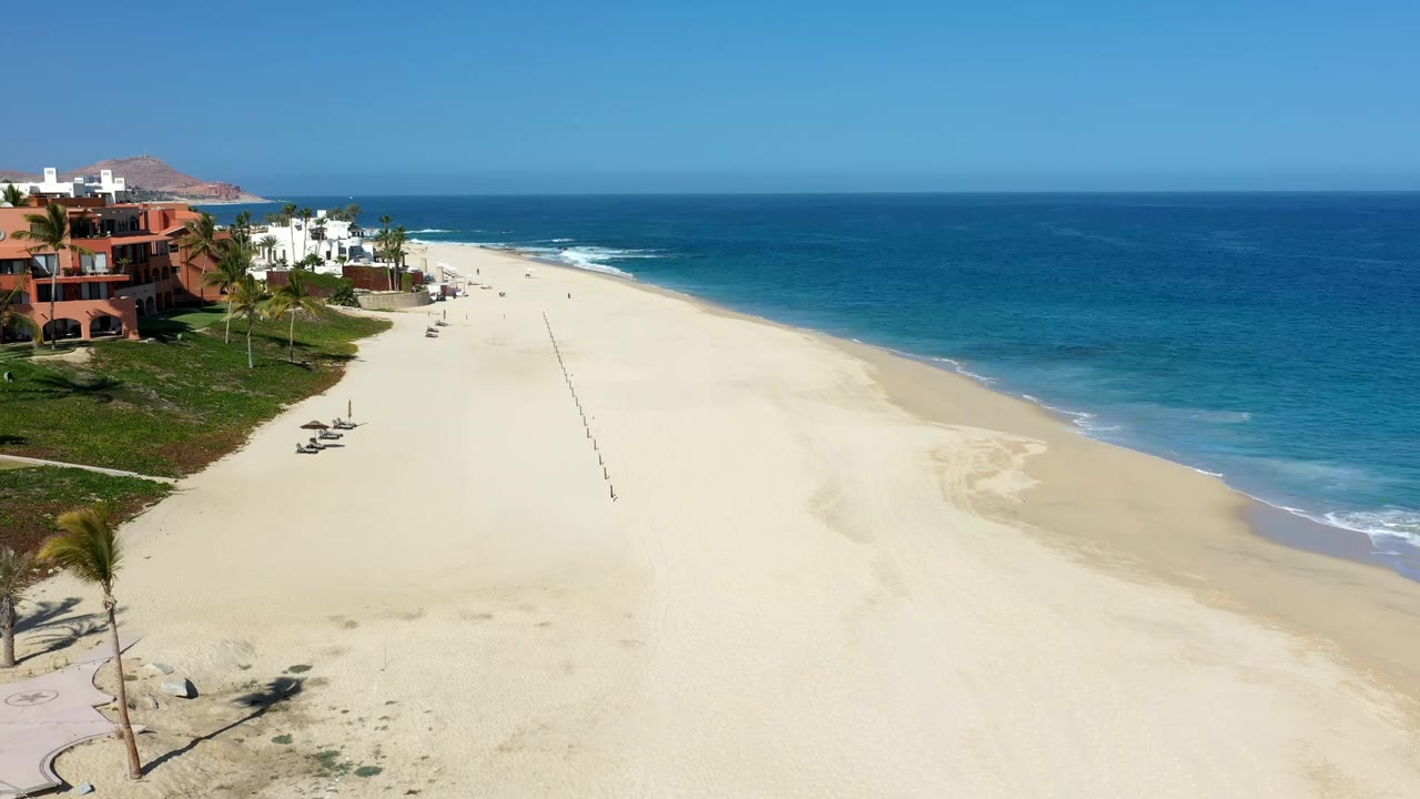 Aerial view in Los Cabos, Mexico, with terracotta and white beachfront houses at left, a broad empty stretch of sand, surf edging the right side, and small people scattered in the distance.