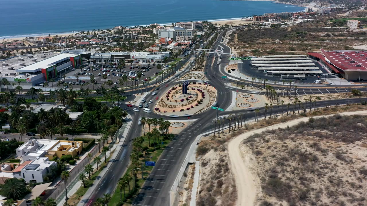 Opening aerial frame over Los Cabos, Mexico showing cars around a landscaped roundabout, palm-lined roads, commercial buildings, dry scrub foreground, ocean horizon, and no visible people.