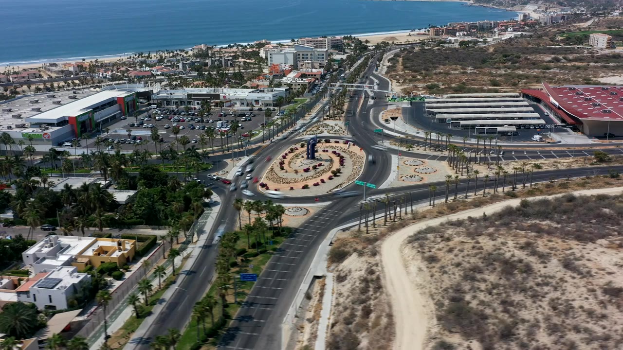 Second aerial frame in Los Cabos, Mexico with the roundabout centered lower in view, cars on multiple approach roads, palms lining the median, beachfront buildings in the distance, and no visible people.