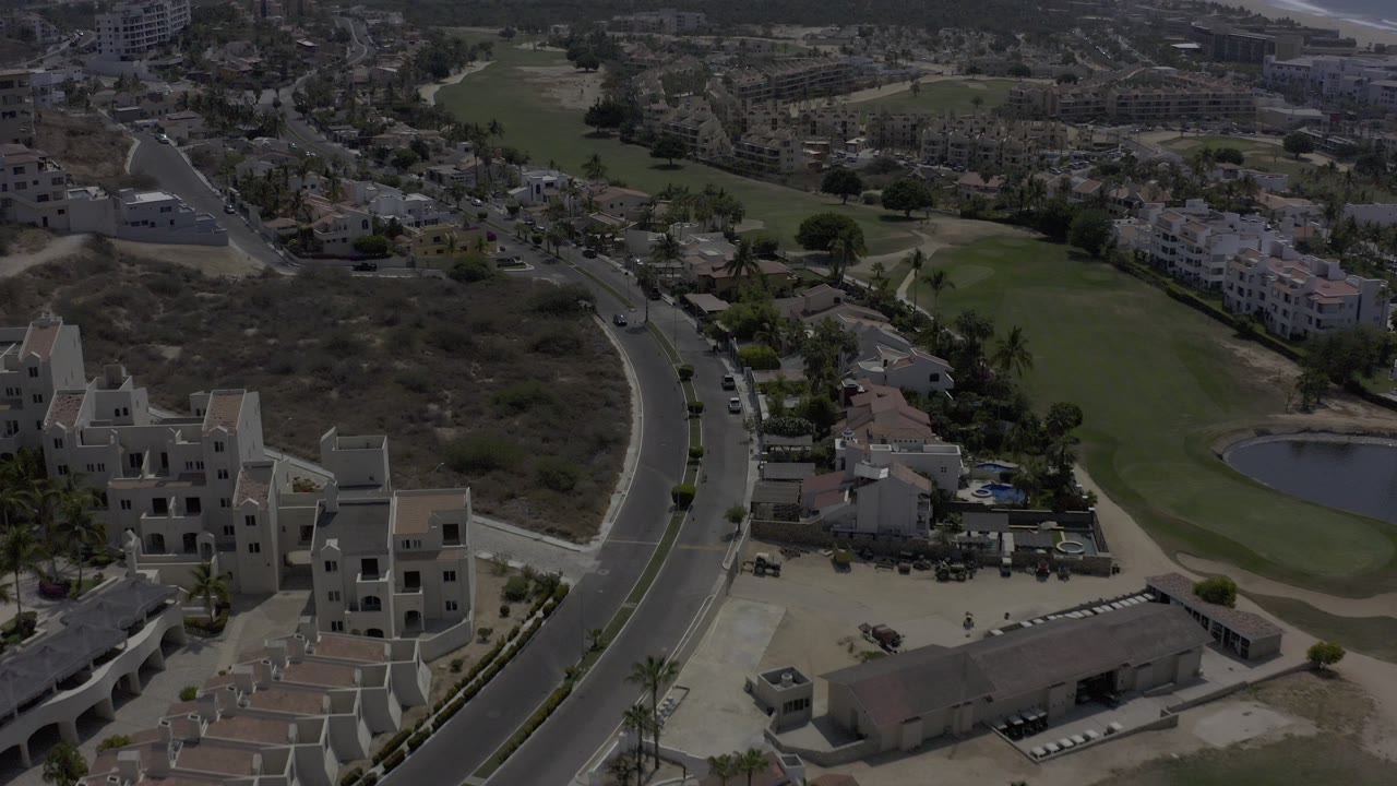 A high aerial frame over Los Cabos shows a curved road beside pale multi-story buildings, brown open lots, and a strip of green golf-course grass, with no recognizable people visible.