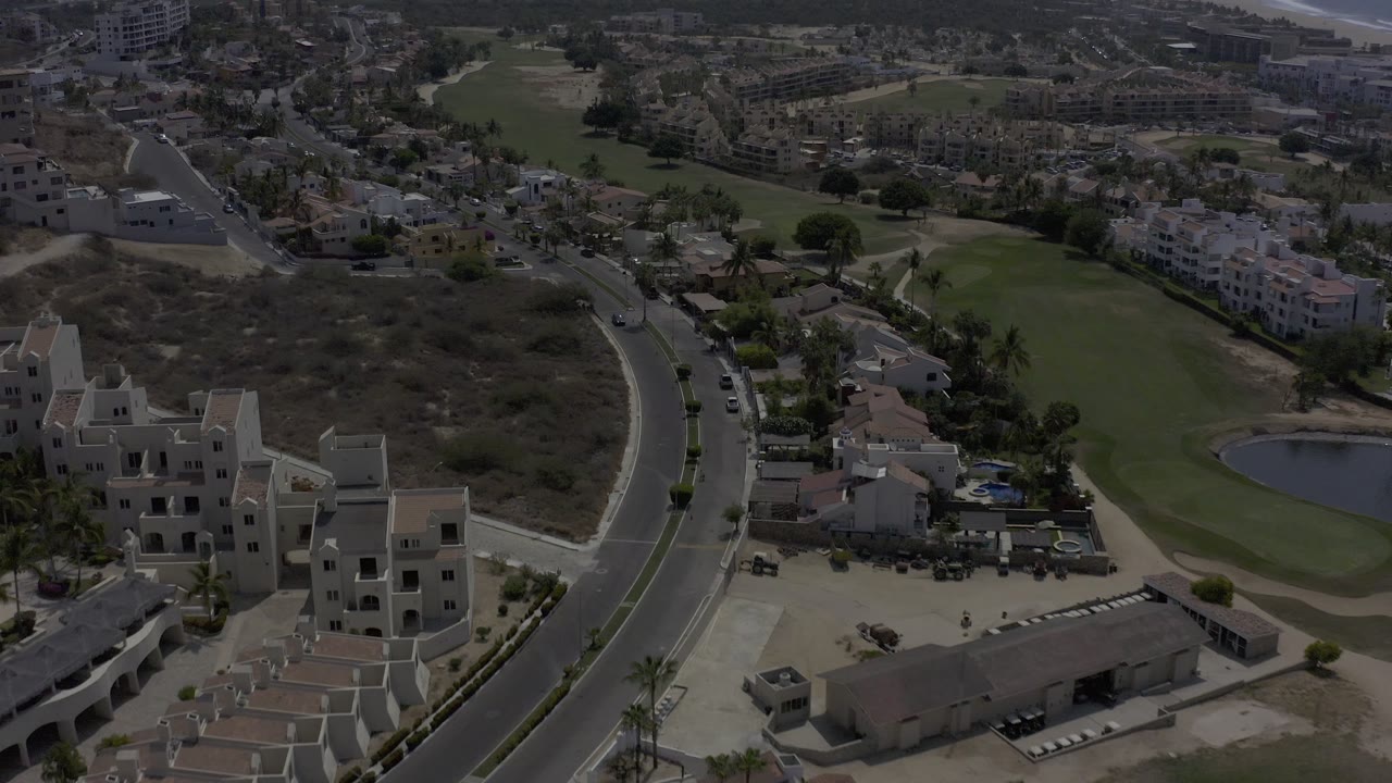 The next Los Cabos aerial view keeps the curving road central while showing cars, pale buildings, nearby homes, dry terrain, green grass, and a small pond, with no recognizable people visible.