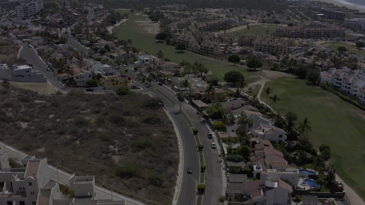 Opening aerial frame over Los Cabos, Mexico, with a curved road, parked cars, private homes, dry scrubland, and green fairway areas, with no visible people.
