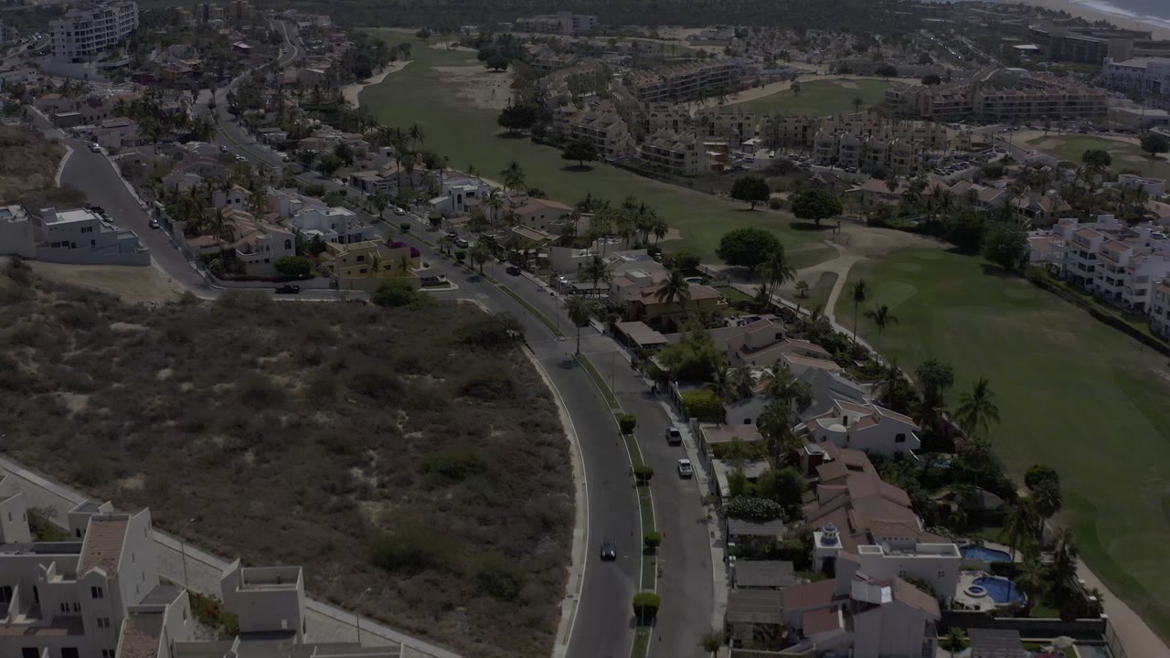 A later aerial view of Los Cabos, Mexico, showing the road running up through homes, palms, dry terrain, green fairways, and clustered buildings in the distance, with no visible people.