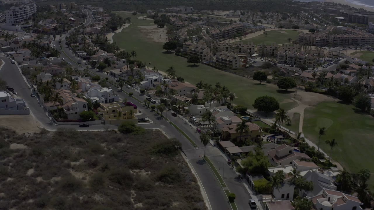 A Los Cabos aerial frame looks across low-rise homes, a road intersection, palms, and a long green landscaped area running through the neighborhood, with no recognizable people visible.