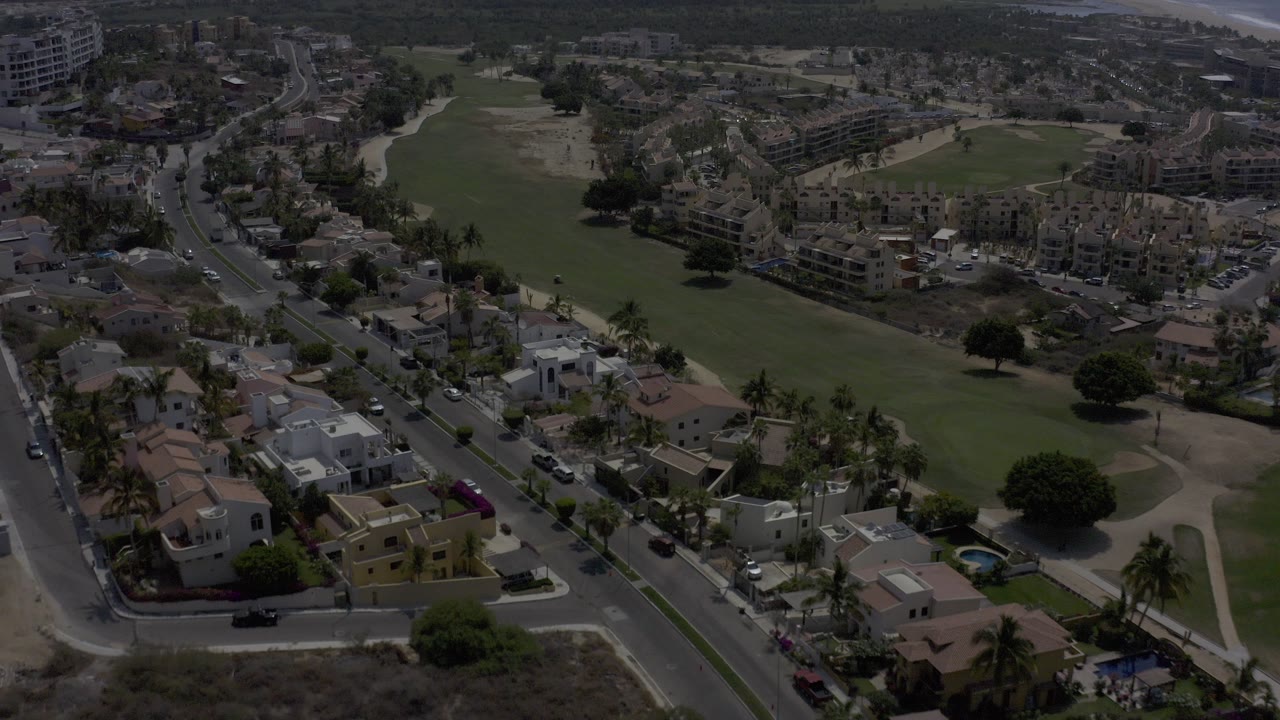 Opening aerial frame over Los Cabos, Mexico, with residential roofs and streets in the foreground, a broad green field beyond them, distant buildings, and no visible people.