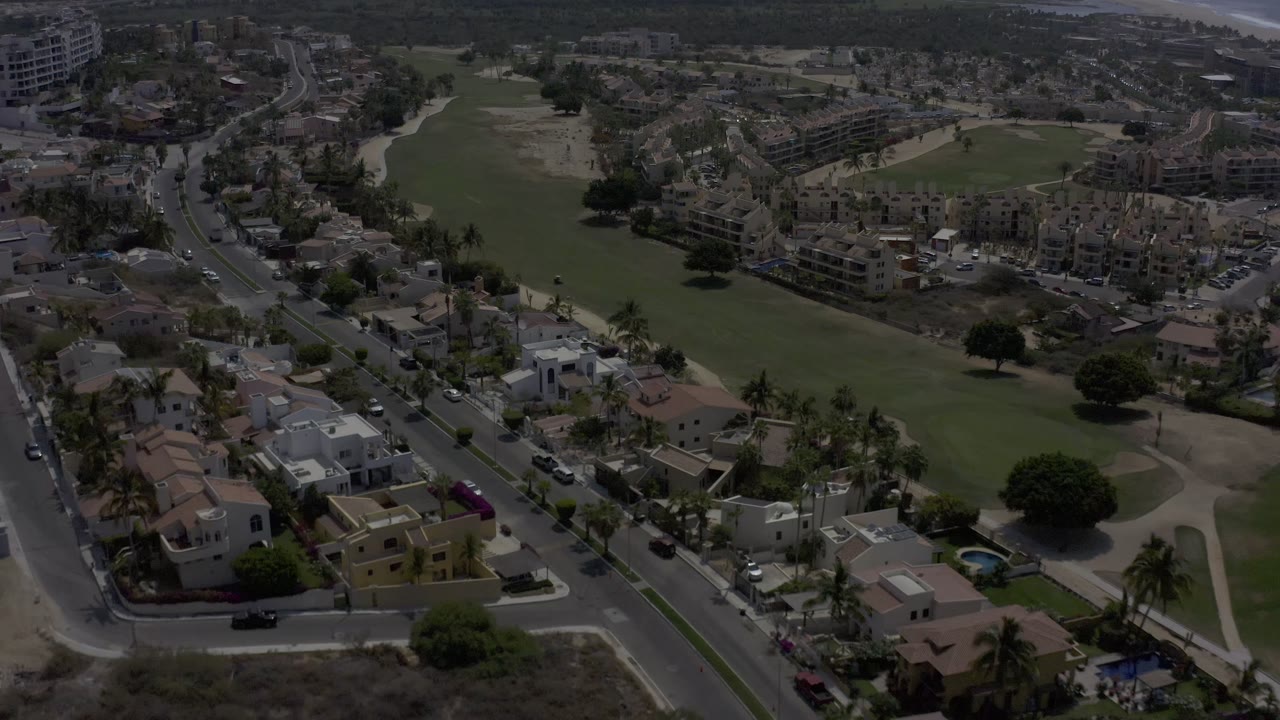 A Los Cabos, Mexico aerial view holds on a green field running past residential blocks, with curving roads, parked vehicles, palm trees, and no visible people.