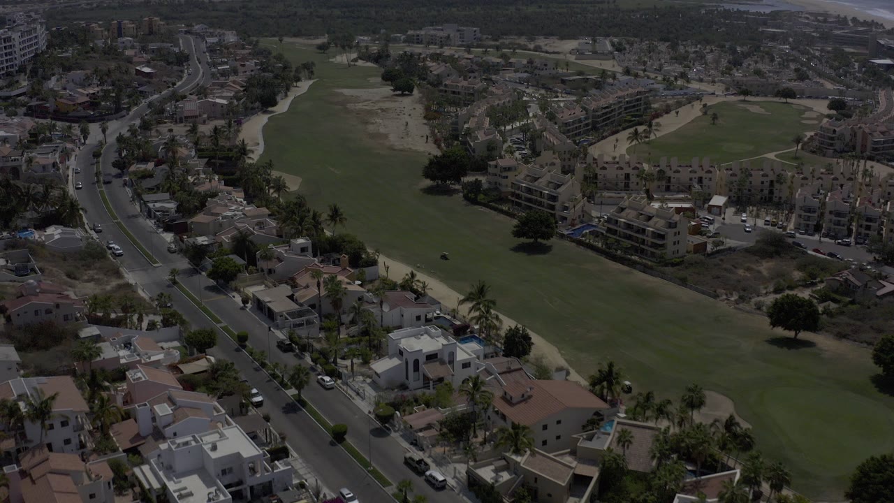 Drone-height view over Los Cabos with a broad green area, palm-lined road, low-rise homes, and distant buildings, with no visible people.