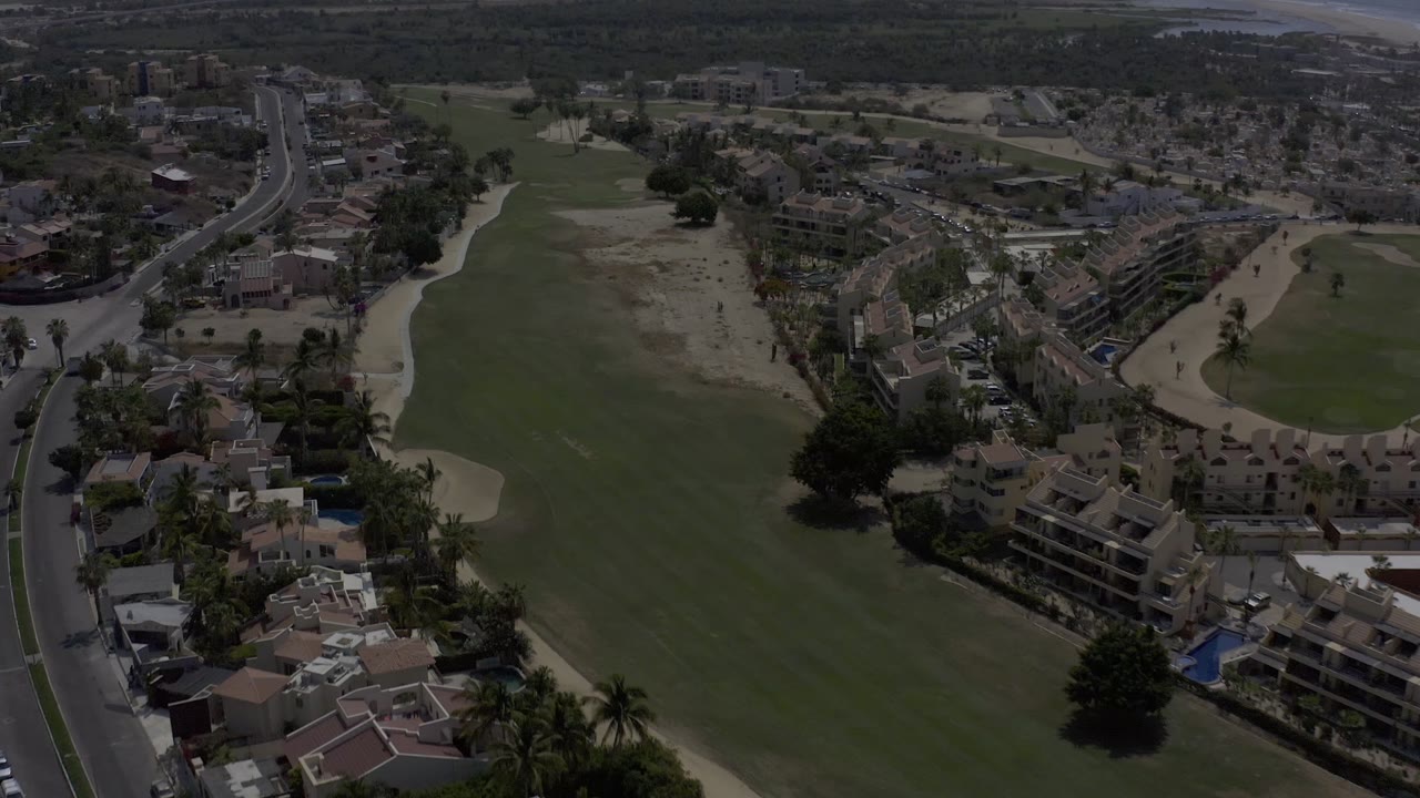 High aerial frame in Los Cabos, Mexico with the fairway centered, a road running up the left side, low-rise homes at the bottom, dry tan areas, and no visible people.