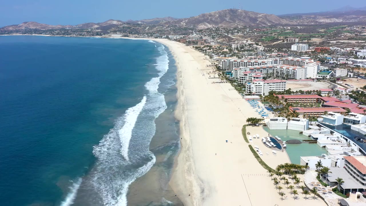The Los Cabos aerial holds the coastline with a larger surf band rolling toward the sand, hotel buildings on the right, ocean filling the left side, hills beyond town, and no visible people.