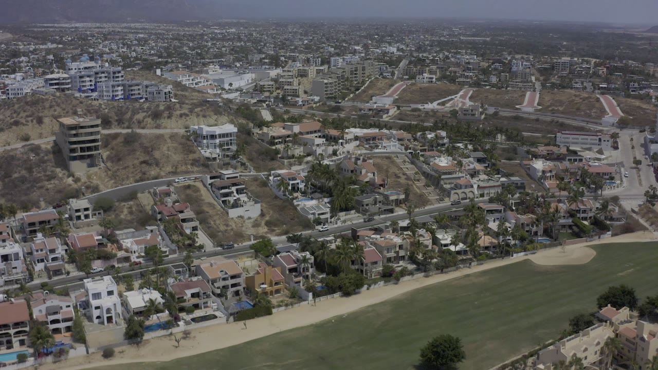 Los Cabos hillside neighborhood seen from above, with villas, terraced roads, dry slopes, a golf fairway in the foreground, and no visible people.