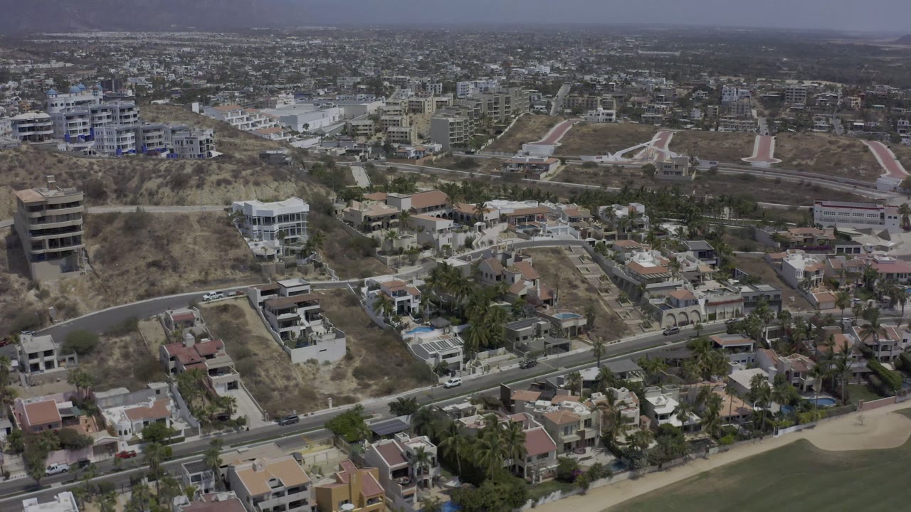 Los Cabos aerial view with villas in the foreground, dry hillside lots, larger modern buildings in the midground, and no visible people.