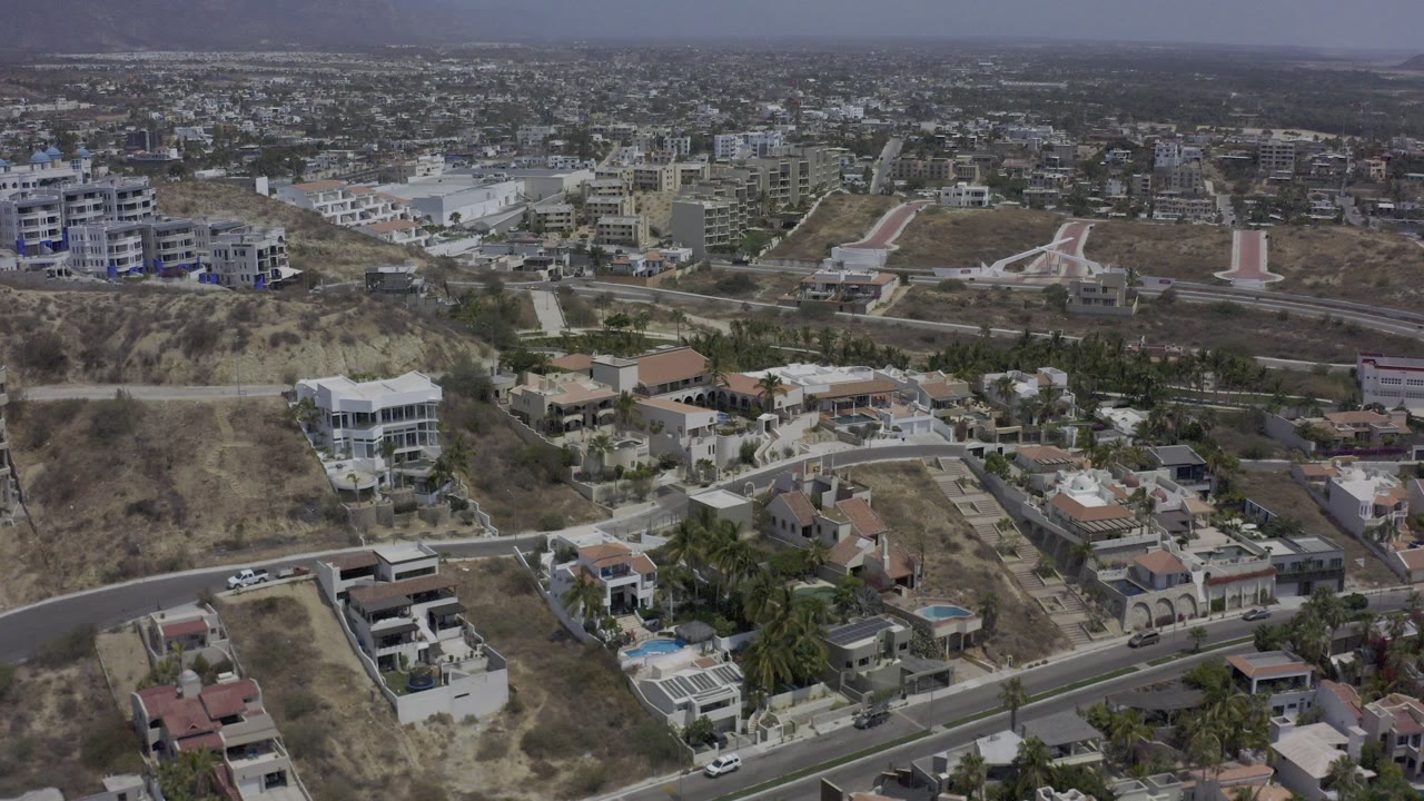 Aerial view over Los Cabos hillside homes with winding roads, dry brown slopes, private pools, dense buildings in the distance, and no visible people.