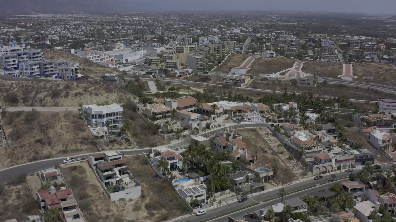 Los Cabos aerial frame with large hillside houses, a curved road in the foreground, pale dry slopes, mid-rise buildings beyond, and no visible people.